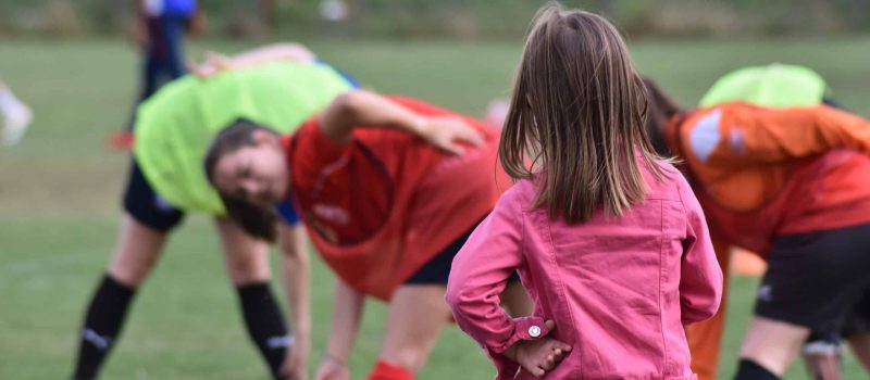 petite fille de dos devant un entrainement de joueuses de football plus âgées